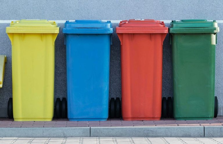 Bunte Abfallbehälter in Gelb, Blau, Rot und Grün nebeneinander auf einem Gehweg.